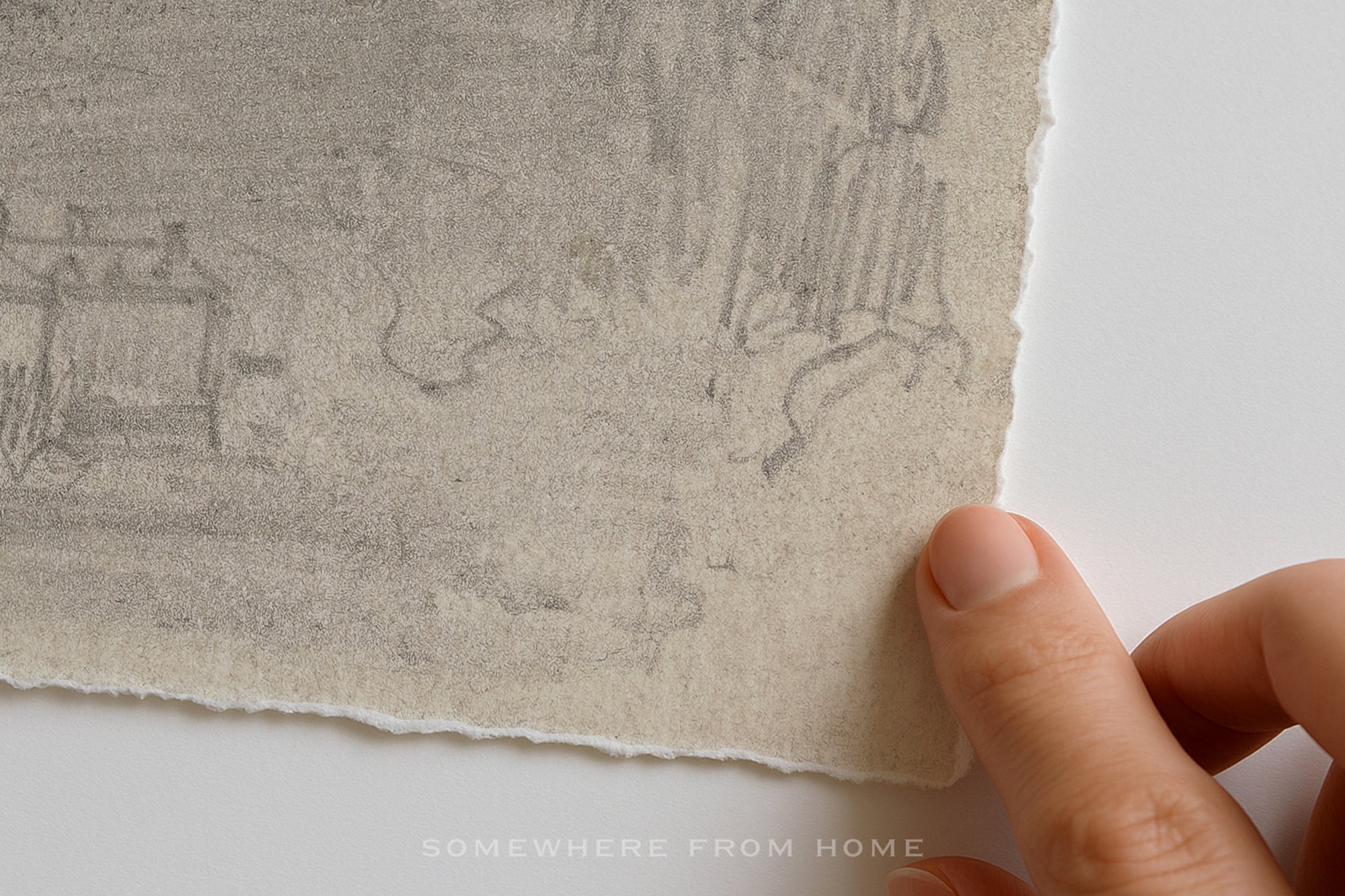 A close-up of a hand holding the edge of a textured handmade paper sheet with a faint pencil sketch, against a plain white background.
