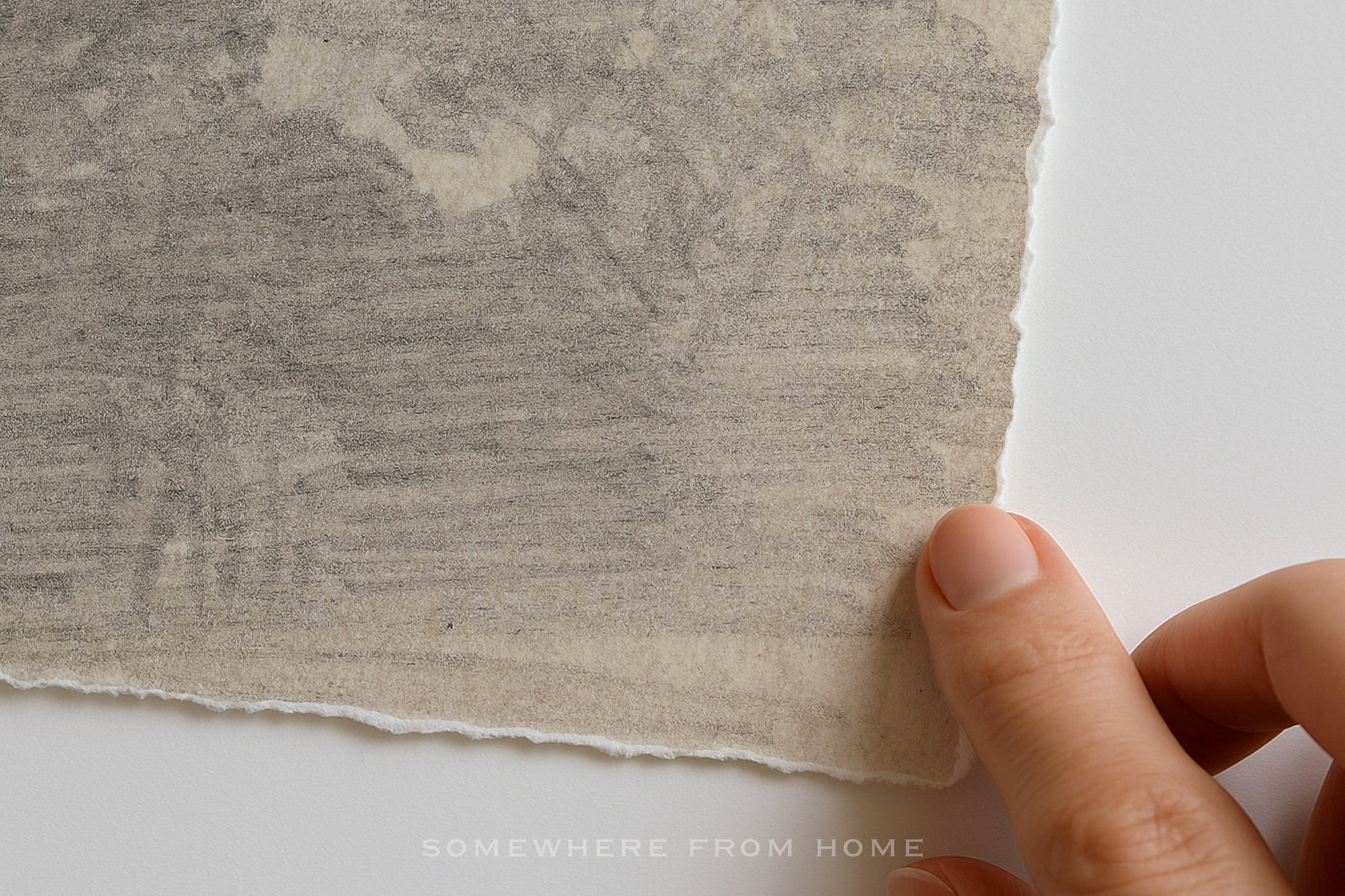 A close-up of a hand holding the edge of a textured, off-white handmade paper sheet with torn margins against a plain white background. A faint watermark at the bottom reads 'SOMEWHERE FROM HOME'.