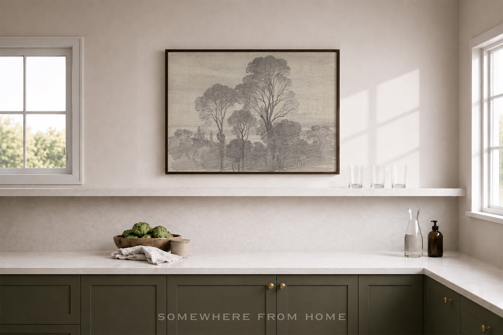 A framed black and white landscape art print hangs on a beige wall above a white shelf in a modern kitchen setting with dark green cabinets and white countertops.