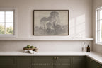 A framed black and white landscape art print hangs on a beige wall above a white shelf in a modern kitchen setting with dark green cabinets and white countertops.
