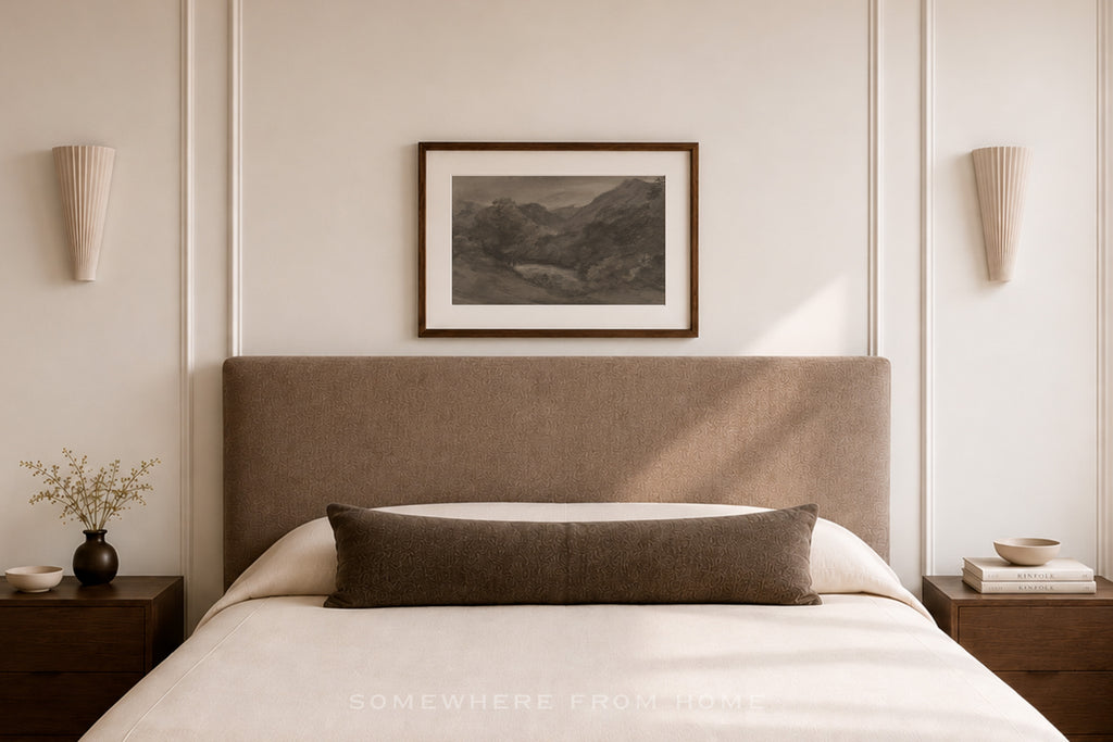 A styled bedroom scene featuring a bed with a tall upholstered headboard, white bedding, a dark brown lumbar pillow, matching bedside tables and sconces, and a framed landscape print on the wall.