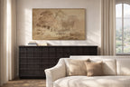 A framed landscape art print in warm beige and brown tones hangs on a cream wall above a dark wood console table, with a light beige sofa in the foreground and a window with curtains on the right.
