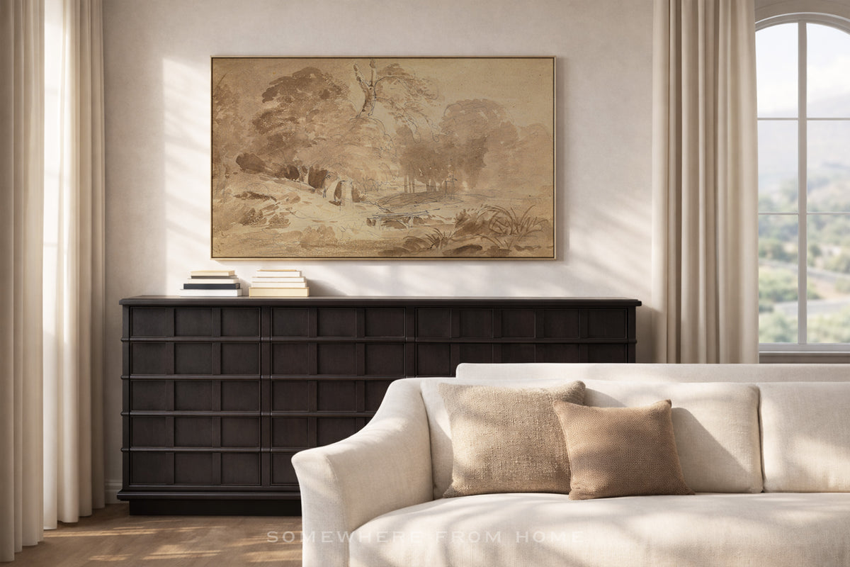 A framed landscape art print in warm beige and brown tones hangs on a cream wall above a dark wood console table, with a light beige sofa in the foreground and a window with curtains on the right.