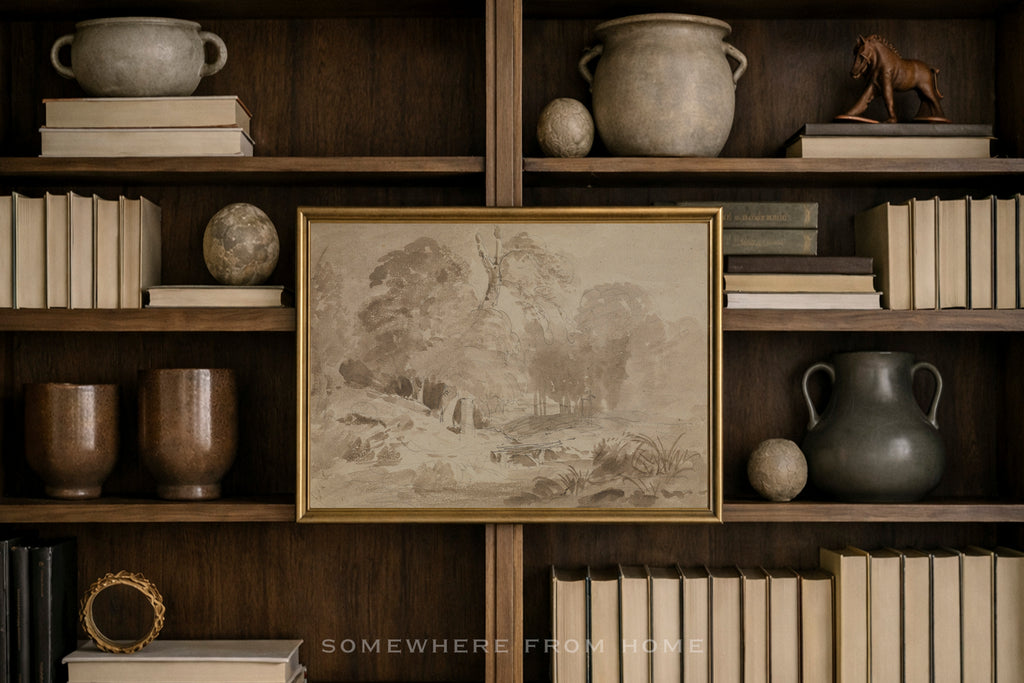 A framed sepia-toned landscape art print with a gold frame, displayed on a dark wood bookshelf surrounded by books, ceramic vases, and decorative objects.