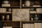 A framed sepia-toned landscape art print with a gold frame, displayed on a dark wood bookshelf surrounded by books, ceramic vases, and decorative objects.