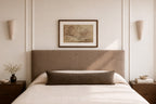 A styled bedroom scene featuring a bed with a tall upholstered headboard, white bedding, a dark brown lumbar pillow, a framed landscape print on the wall, and matching wall sconces.
