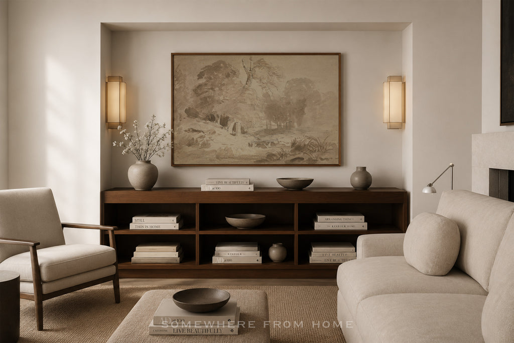 A styled living room interior featuring a large framed landscape painting on a beige wall above a dark wood console. The room includes a light beige sofa, a matching armchair, a coffee table, and decorative ceramic vases and books.