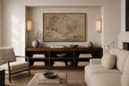 A styled living room interior featuring a large framed landscape painting on a beige wall above a dark wood console. The room includes a light beige sofa, a matching armchair, a coffee table, and decorative ceramic vases and books.