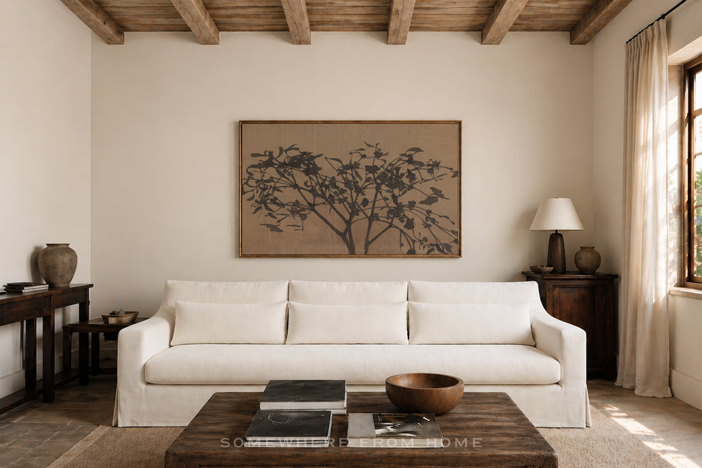 A large framed art print featuring a black ink-style tree on a warm beige background, displayed on a cream-colored wall above a white sofa in a rustic, sunlit living room.