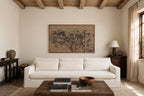 A large framed art print featuring a black ink-style tree on a warm beige background, displayed on a cream-colored wall above a white sofa in a rustic, sunlit living room.