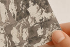 A close-up of a hand holding a square art print or card featuring a monochrome ink-style illustration of a figure and animals on a rocky cliffside. The print has a warm off-white background with gray and black brushwork.