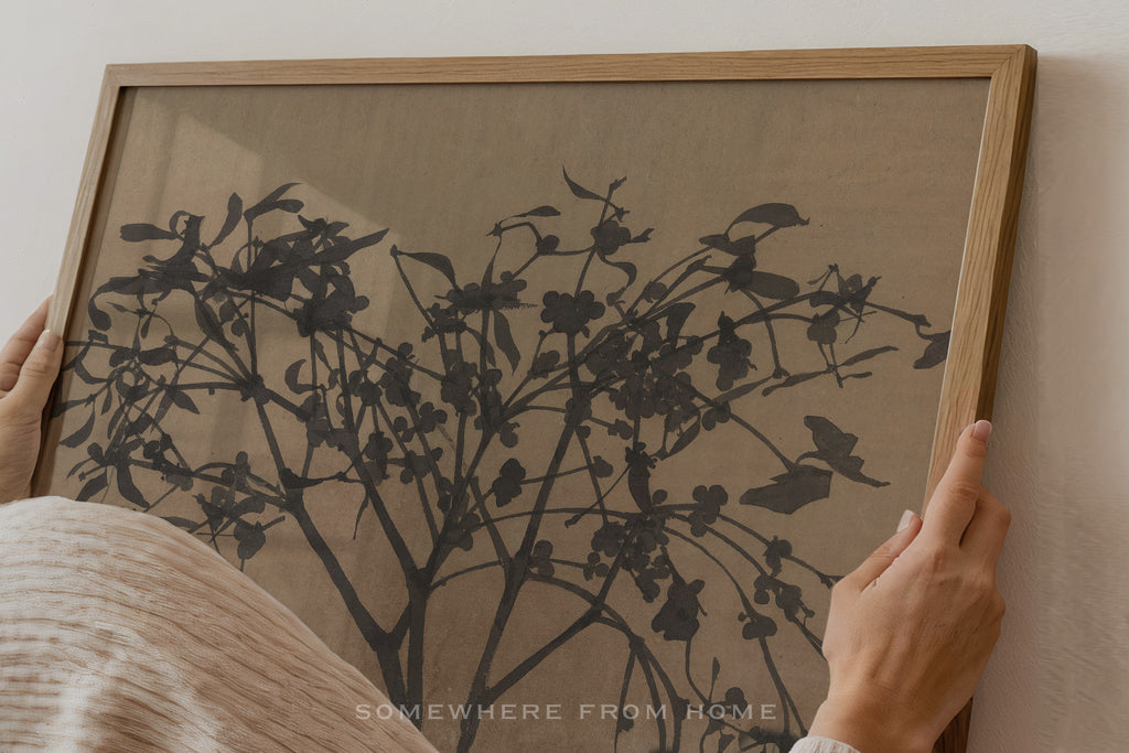 A person's hands are shown holding a light oak wood framed art print featuring a black ink-style botanical tree design on a textured beige background.