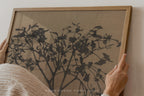 A person's hands are shown holding a light oak wood framed art print featuring a black ink-style botanical tree design on a textured beige background.