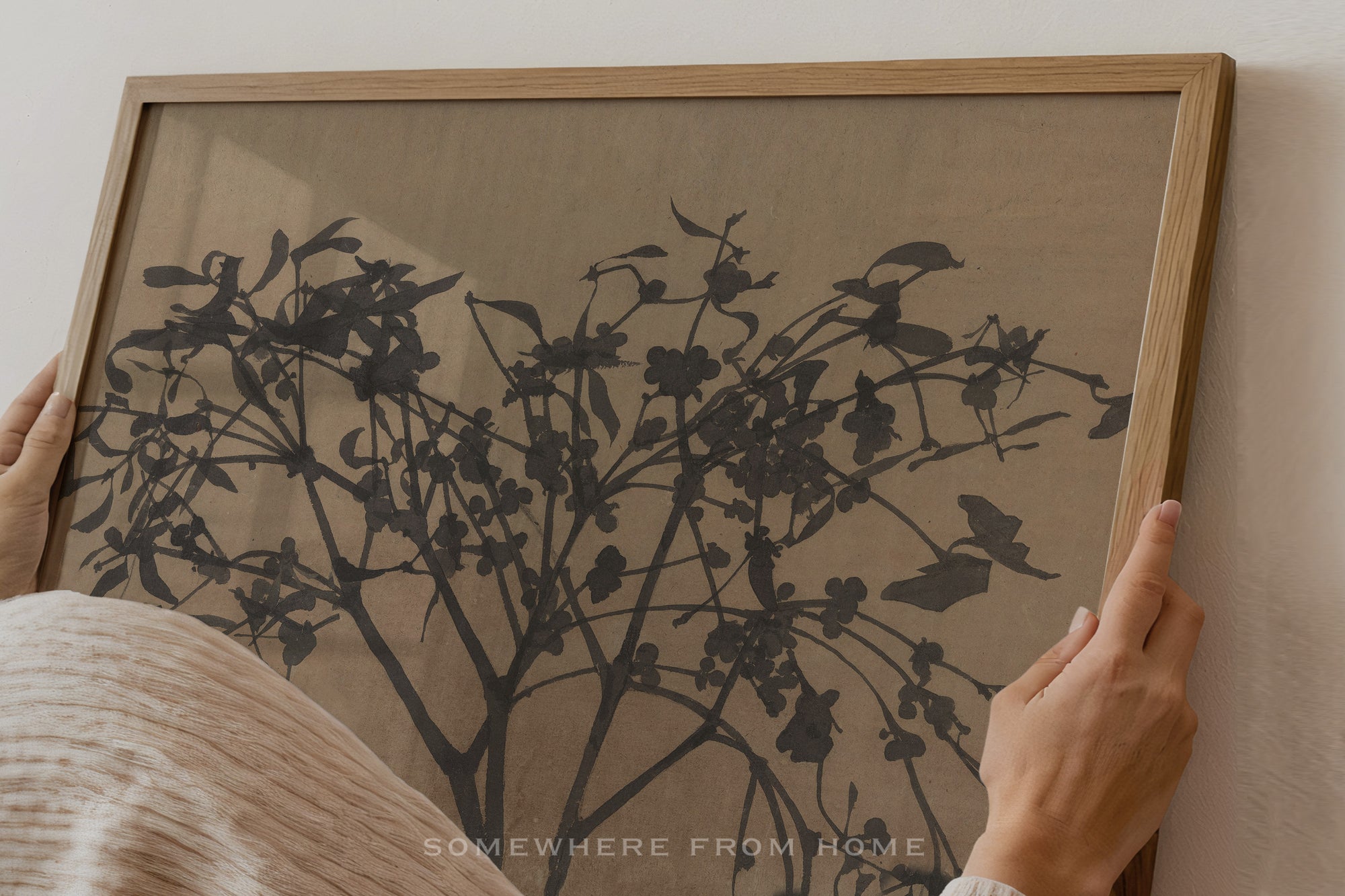 A person's hands are shown holding a light oak wood framed art print featuring a black ink-style botanical tree design on a textured beige background.