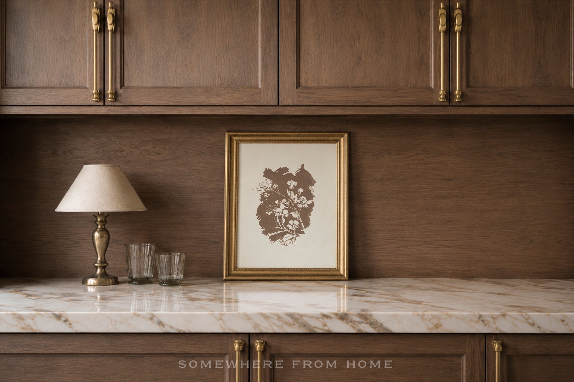 A framed botanical art print with a brown floral illustration on a cream background, styled in a gold frame on a white marble countertop against a dark wood cabinet wall.
